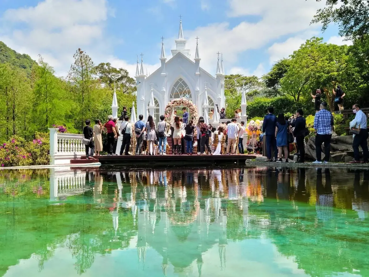 湖畔教堂全景 真愛桃花源白色哥德式教堂與湖畔證婚區全景,賓客立於水岸平台,翠綠湖水完整倒映教堂、山林與藍天白雲