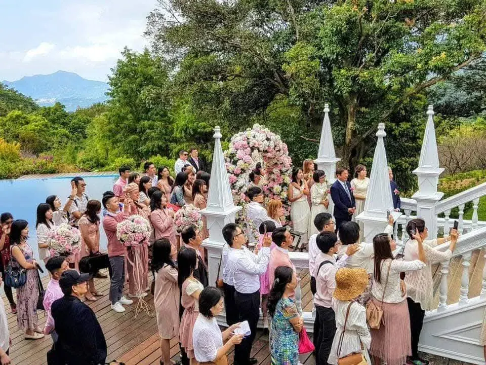 戶外觀禮 真愛桃花源戶外證婚儀式進行中,賓客站在木棧道兩側舉手機拍攝,前方白色尖塔與粉色花球拱門,遠處為陽明山稜線