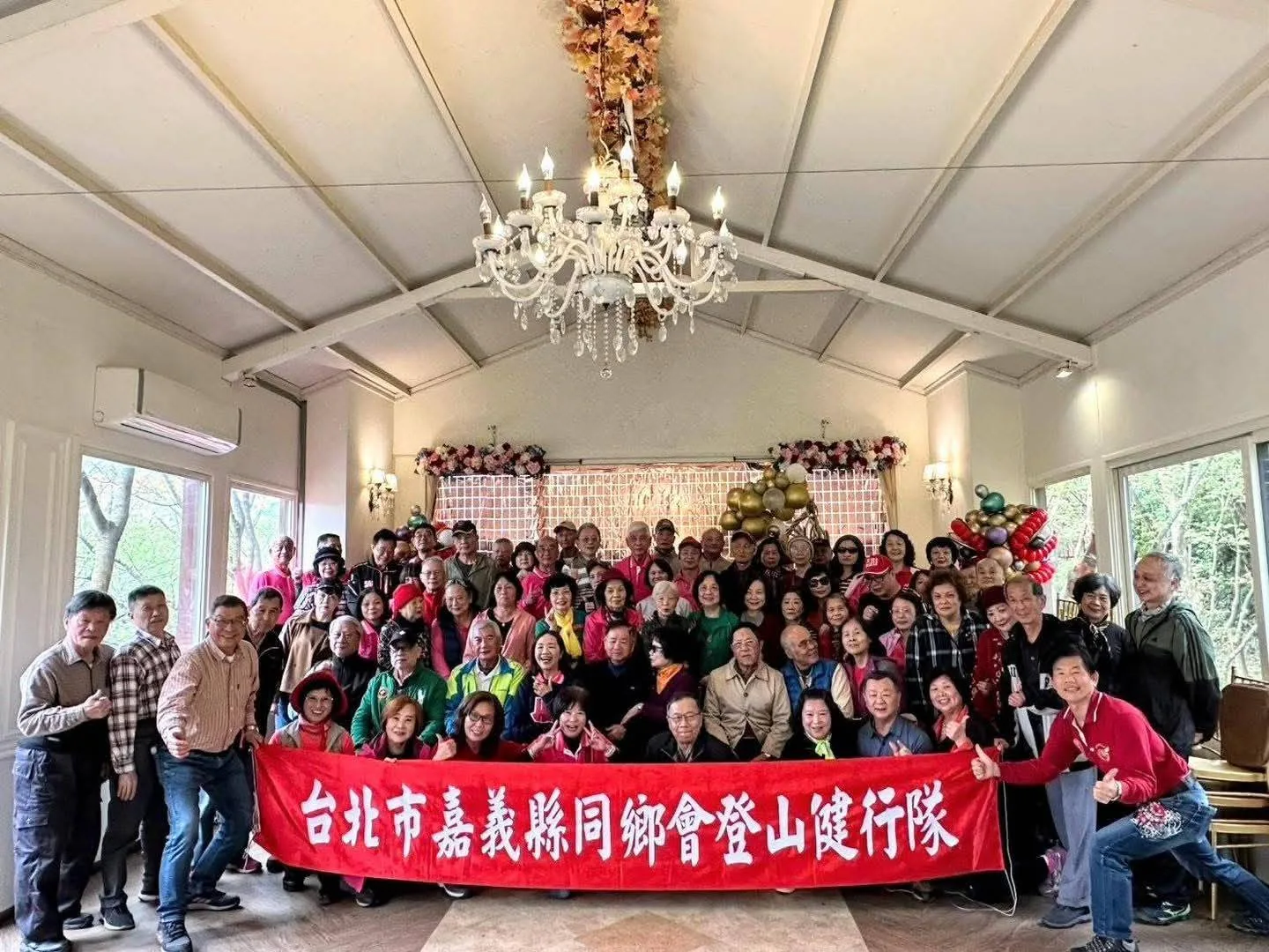 團體包場合照 團體於真愛桃花源室內廳堂包場活動的大合照,全體成員在水晶吊燈與花藝氣球背板前合影,兩側整面採光窗可見園區樹林